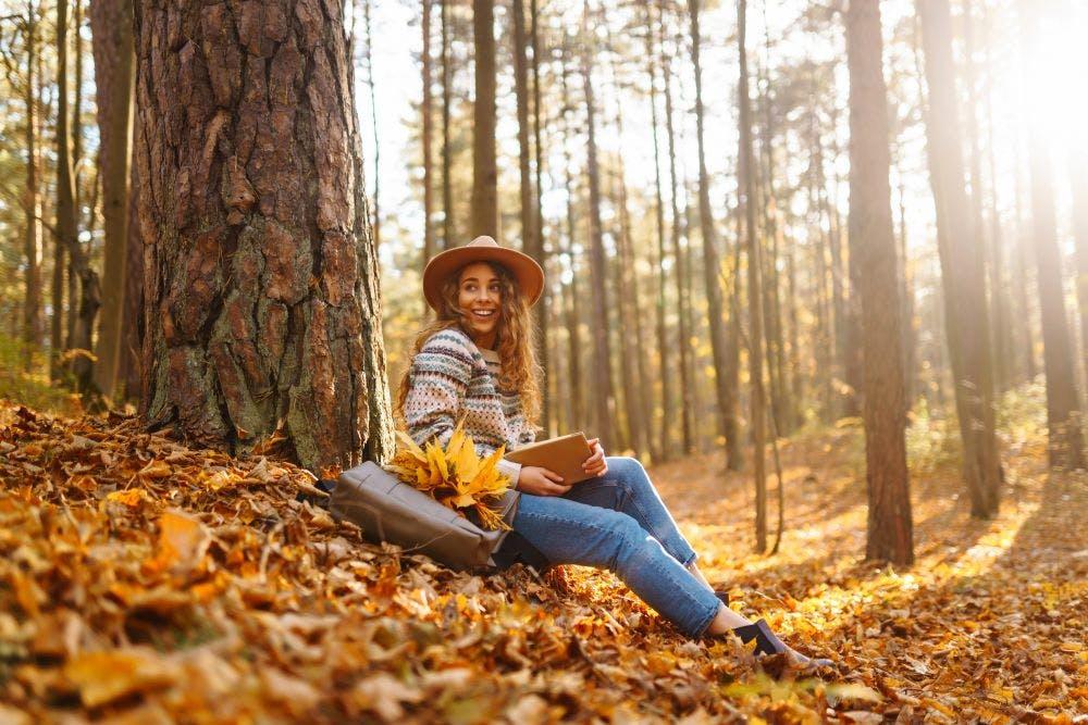 Beautiful smiling young woman student sitting and reading book at autumn park fall yellow foliage leaves.